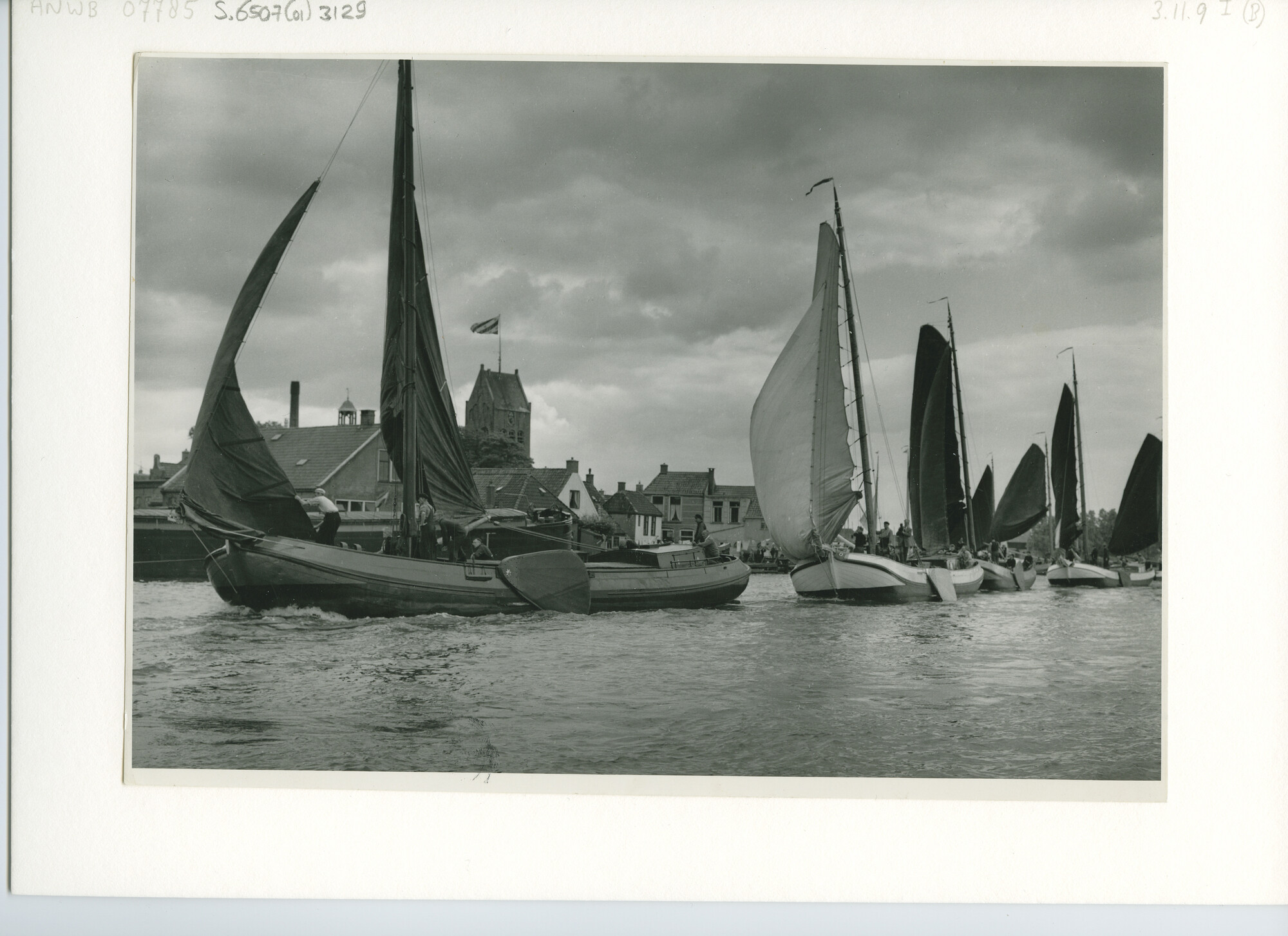 S.6507(01)3129; Vooraangezicht over bakboord van zeven skûtsjes - tjalken - die tijdens Skûtsjesilen achter elkaar in linie varend Grouw passeren; foto