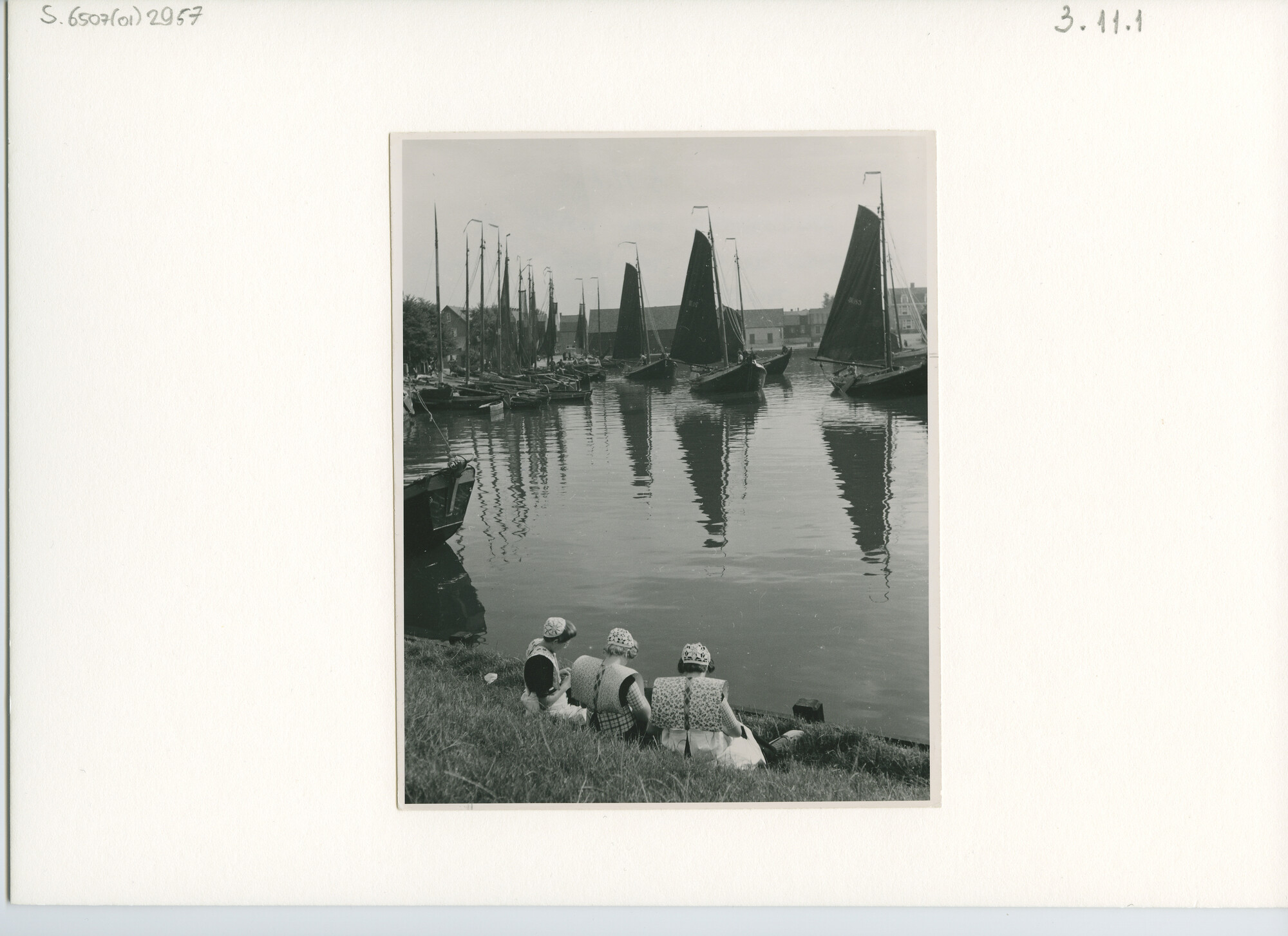 S.6507(01)2957; Botters zeilen onder grootzeil de haven van Spakenburg uit terwijl drie vrouwen in de dracht toekijken; foto