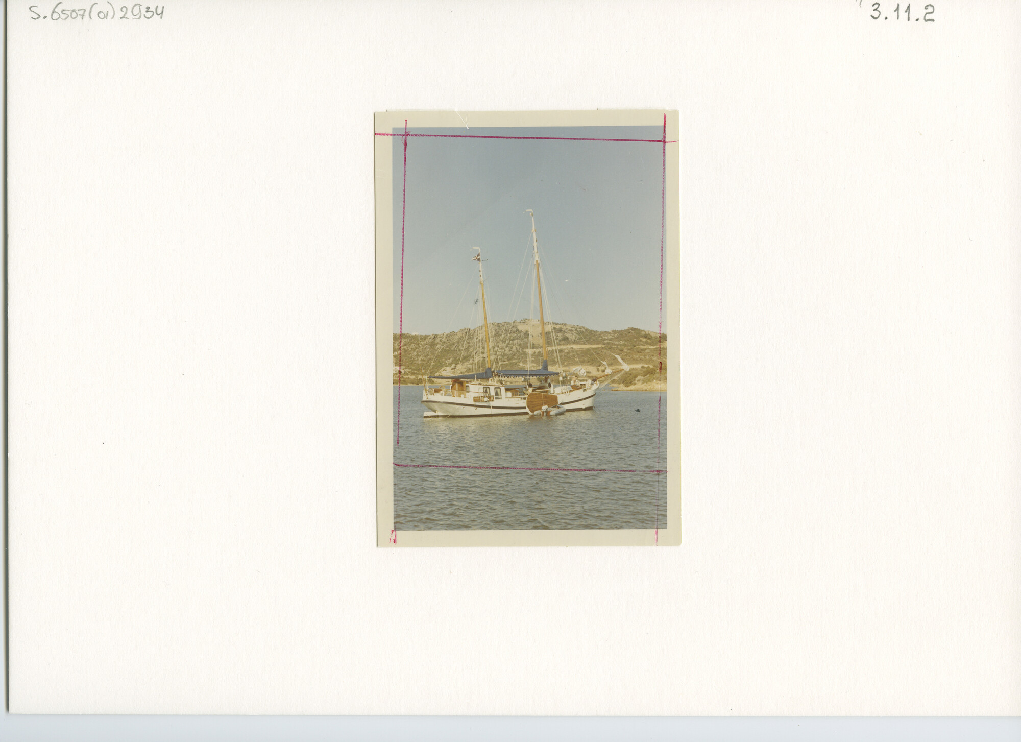 S.6507(01)2934; Een witgeschilderde tweemast klipper met zonnetent voor anker voor een rotsachtig landschap [Middellandse zee].; foto