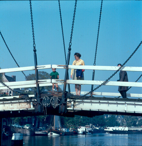 S.9100(5066); Dia van de Amstel met de Magere Brug (detail) met drie mensen erop; diapositief