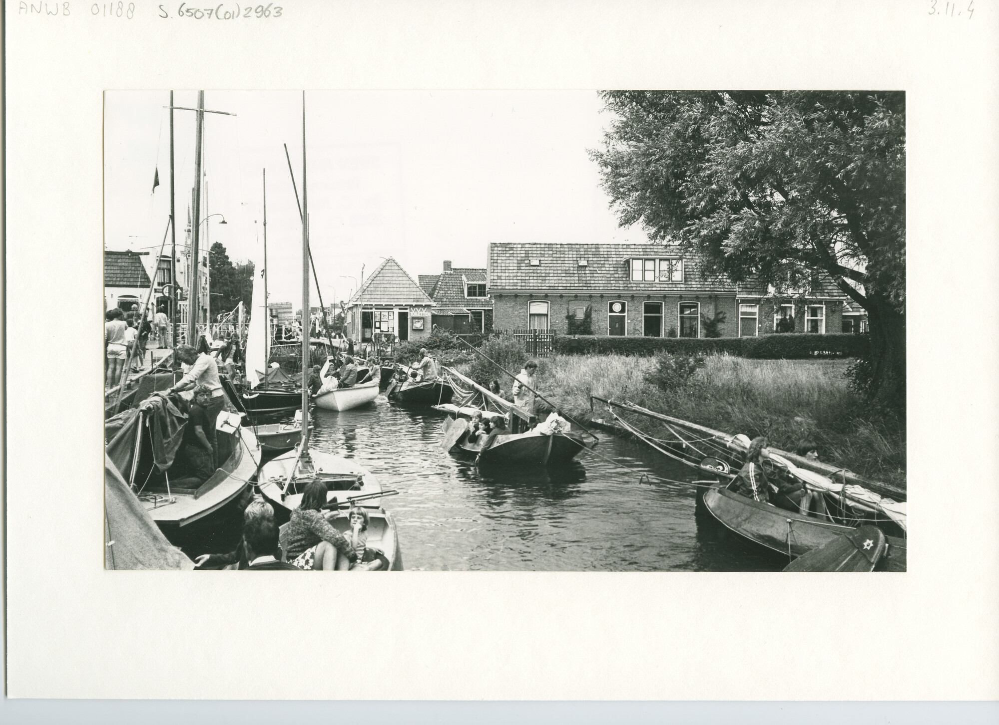 S.6507(01)2963; Een sleep tjotters passeert een aantal zeiljachtjes in het dorp Heeg met het kantoortje van de VVV op de achtergrond; foto